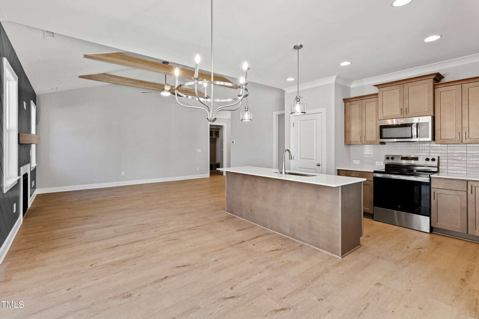 Open-concept kitchen and living area with wood floors, an island with sink, stainless steel appliances, brown cabinets, white countertops, a modern chandelier, and light gray walls. Ceiling beams are visible in the living area.