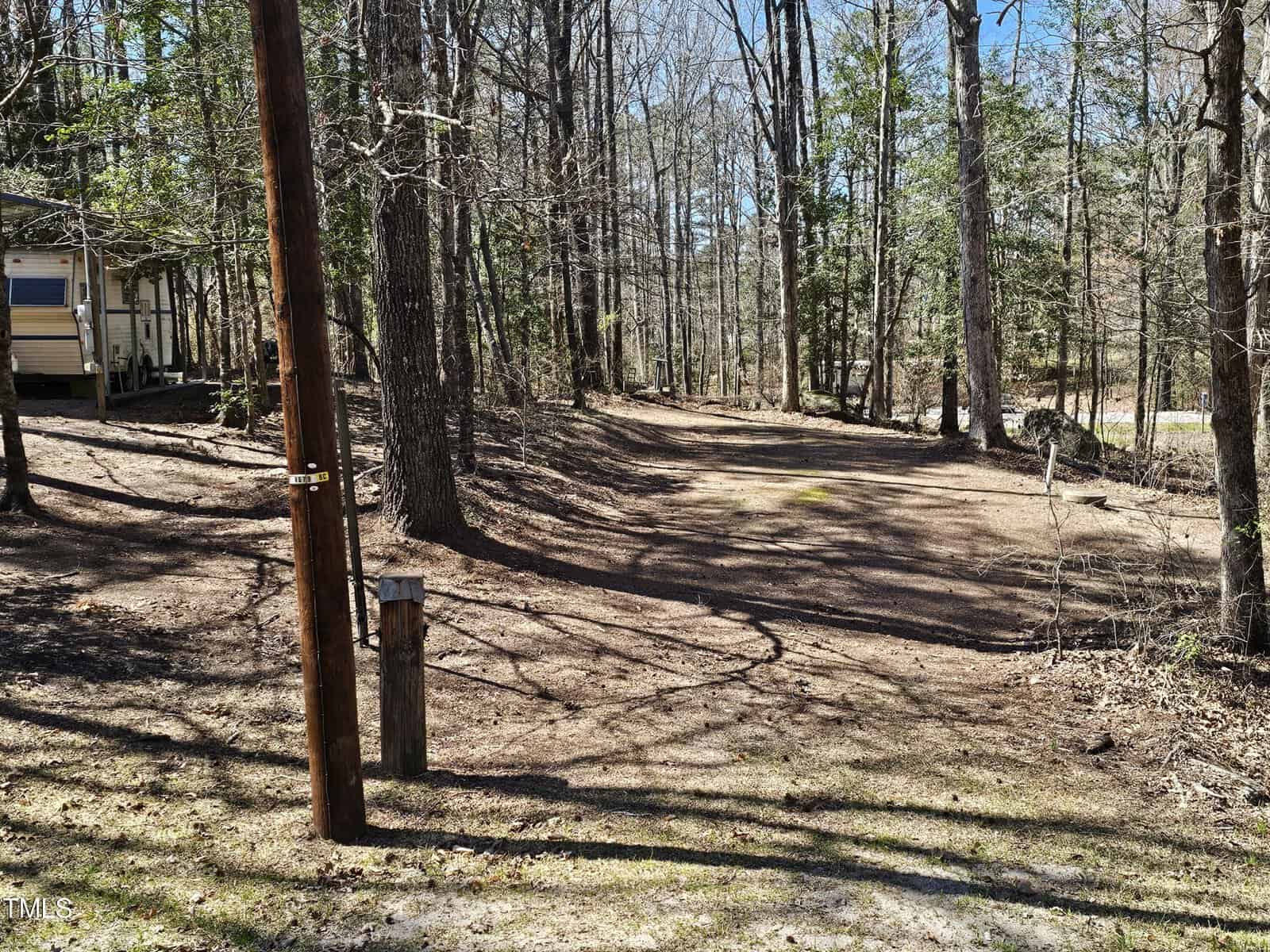 A dirt driveway curves through a wooded area with tall trees. A wooden utility pole stands in the foreground, and part of a beige house or trailer is visible on the left side. Sunlight filters through the trees.