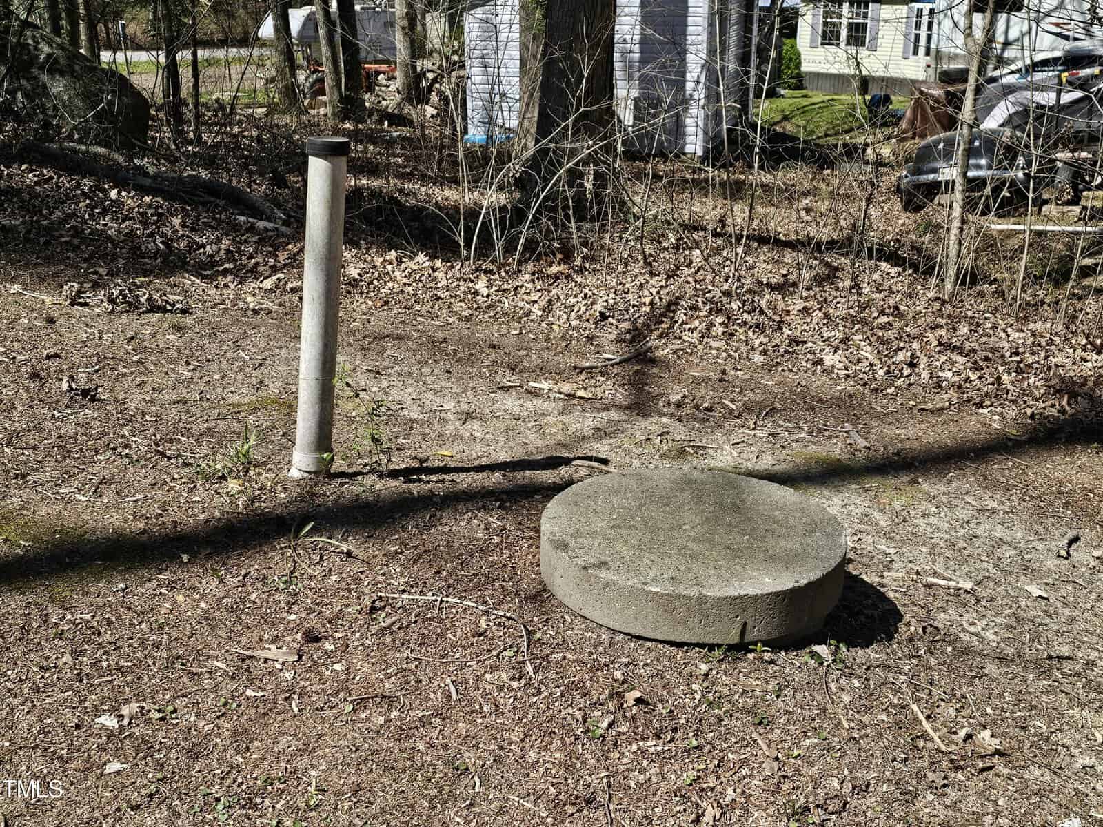 A concrete well cap sits on the ground near a vertical metal pipe in a yard covered with dry leaves; trees, a white shed, and a car are visible in the background.