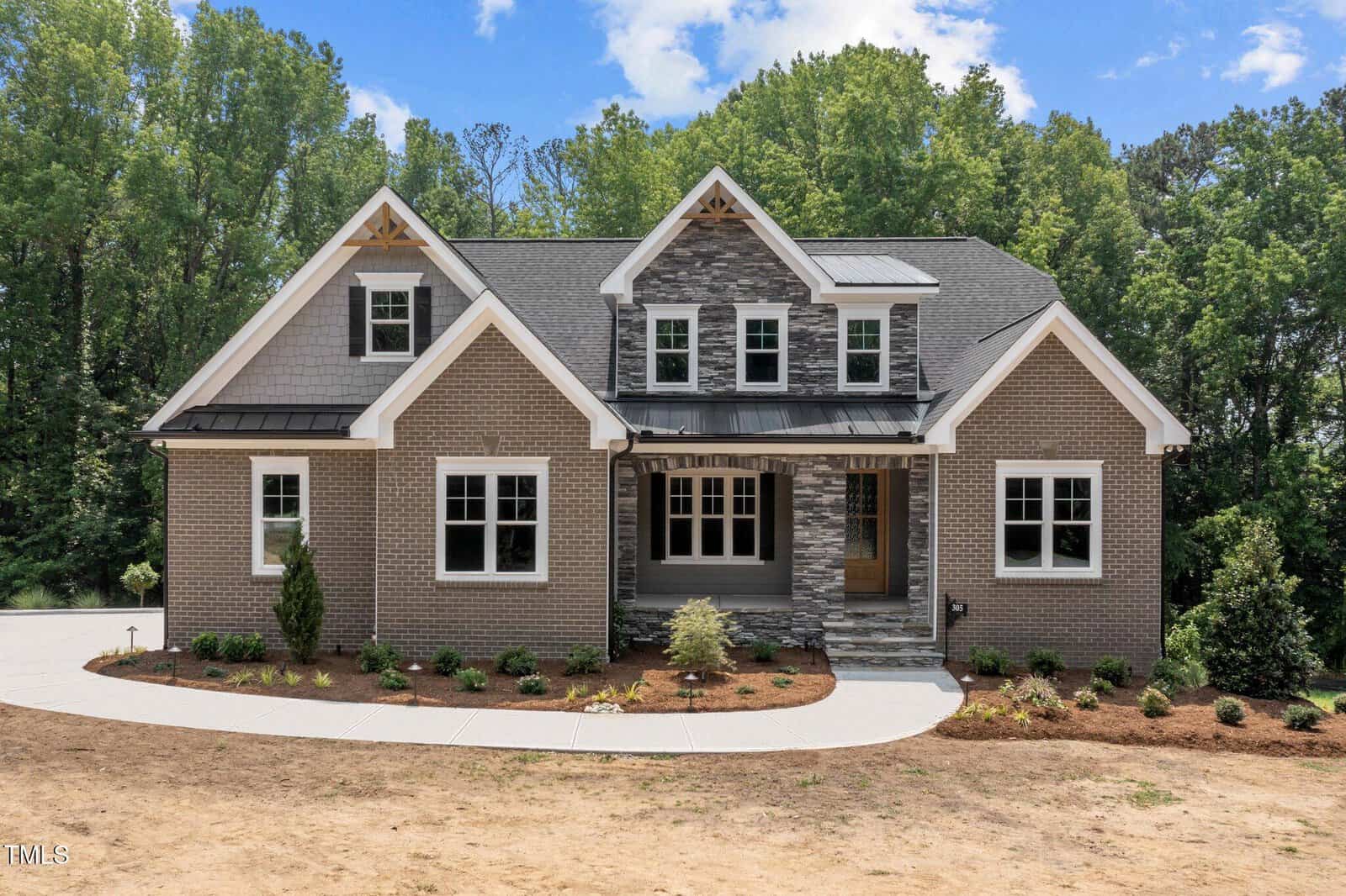 A modern two-story brick house with gray accents, peaked gables, large windows, a covered front porch with stone columns, and landscaped front yard, surrounded by tall green trees under a blue sky with clouds.