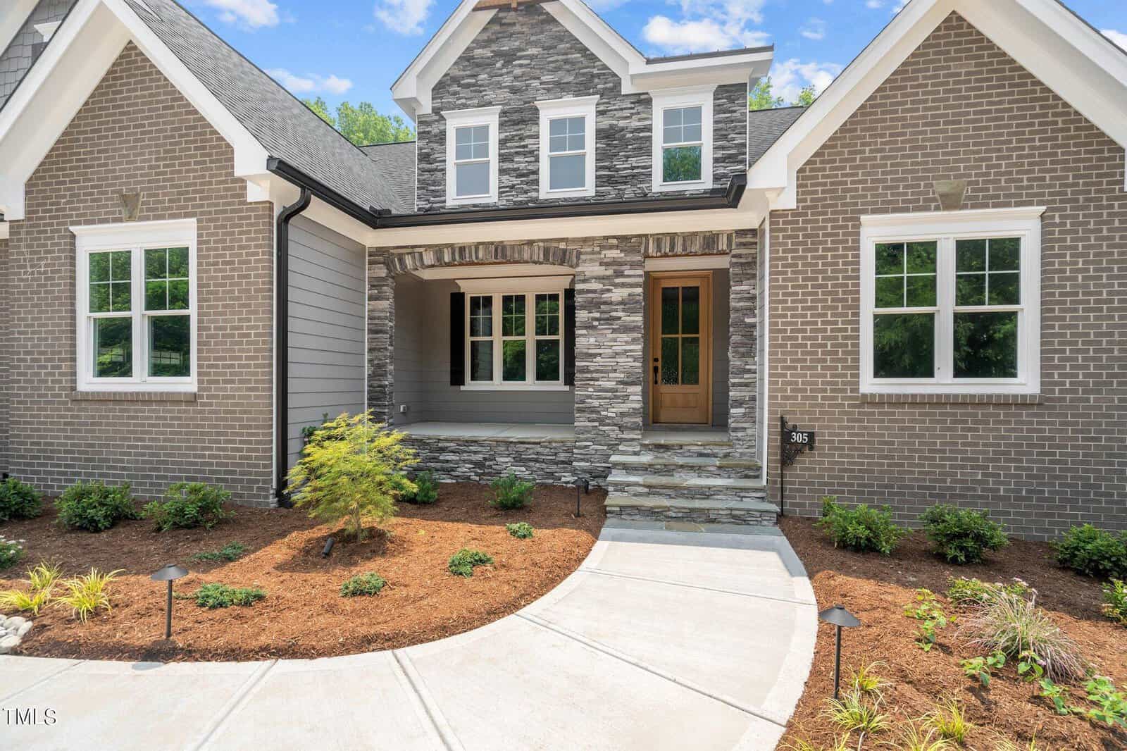 A modern suburban house with brick and stone exterior, a covered front porch, wooden front door, and landscaped yard with mulch and small plants along a curved concrete walkway.