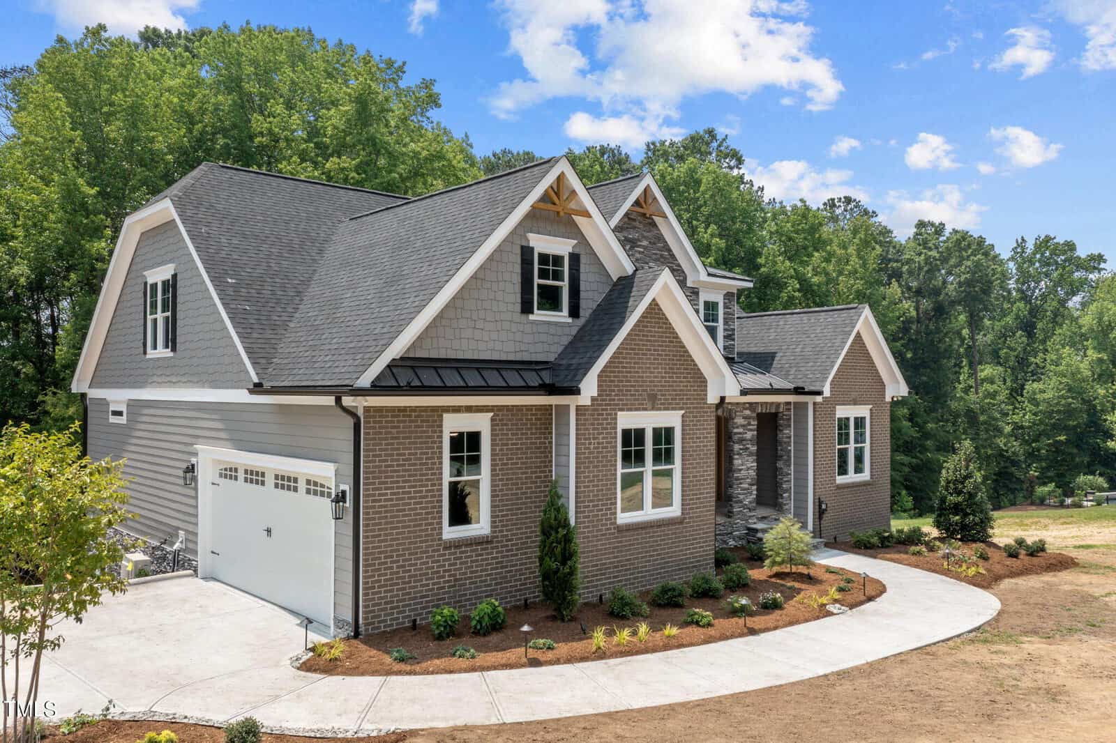 A modern two-story house with gray brick and siding exterior, white trim, and gabled roofs. It has a double garage, neat landscaping, a stone walkway, and is surrounded by trees under a partly cloudy sky.