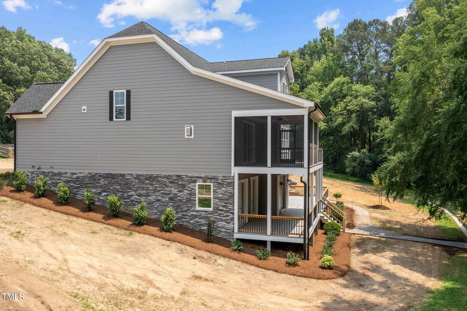 A two-story gray house with stone accents and a sloped yard, featuring a screened-in porch, covered lower deck, and landscaped bushes along the side, surrounded by trees and under a partly cloudy sky.