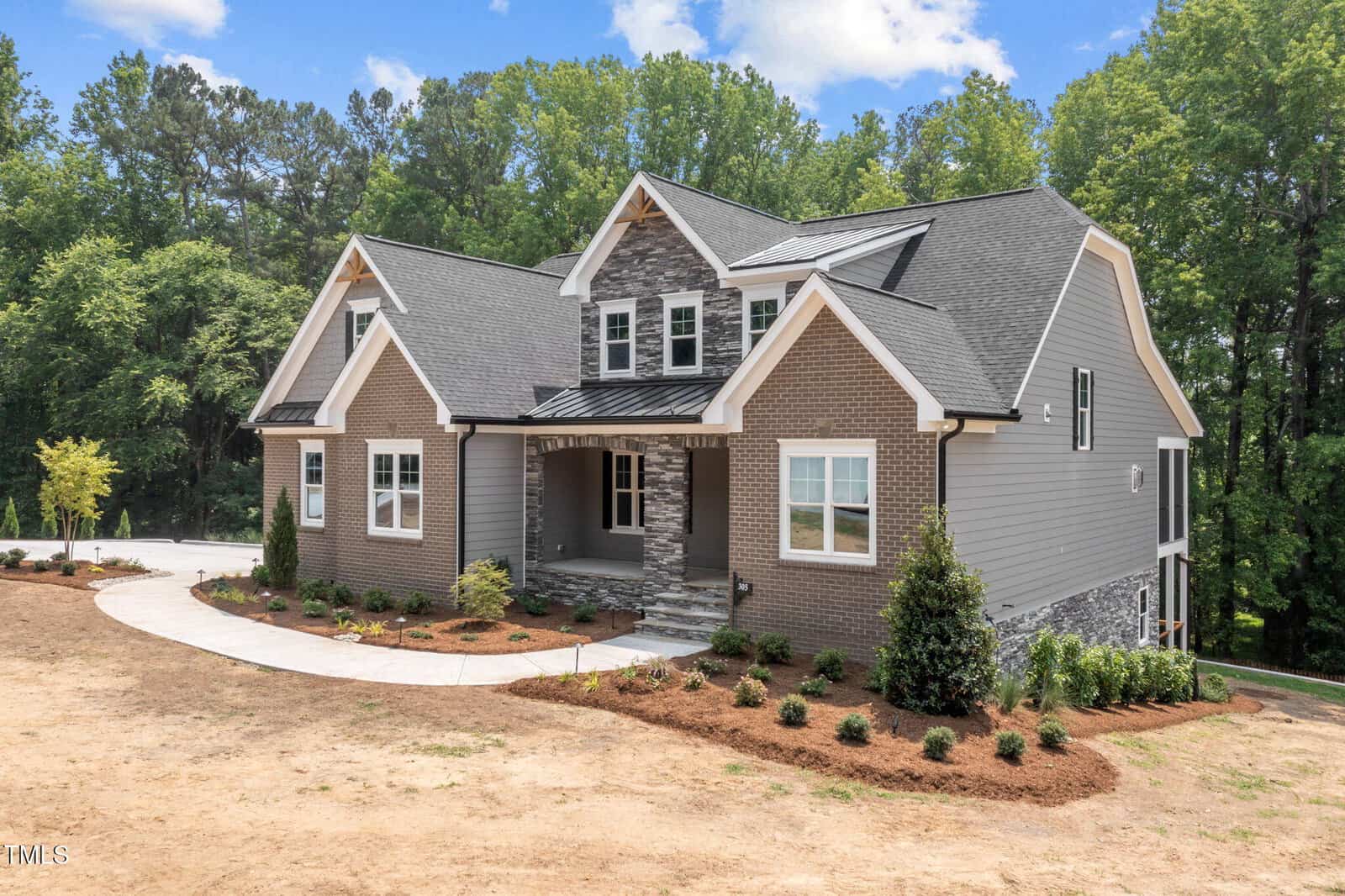 Two-story modern house with gray brick and siding, white trim, gabled roof, and stone accents. The home has a covered front porch, landscaped yard with mulch, and is surrounded by trees under a partly cloudy sky.