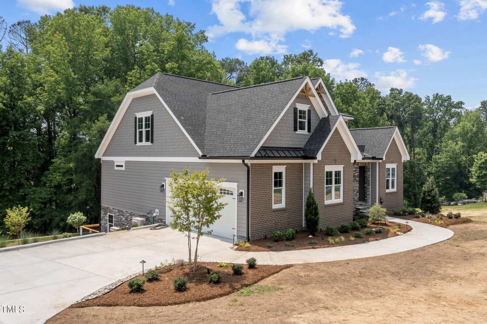 A modern gray house with white trim and a steep roof sits on a landscaped lot with young trees and shrubs, a concrete driveway, and a backdrop of tall green trees under a partly cloudy sky.