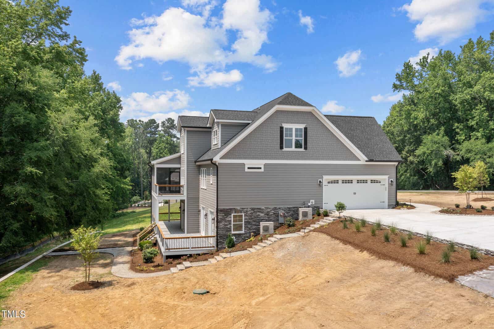 A modern gray two-story house with a covered porch, stone accents, and a three-car garage sits on a newly landscaped lot with a concrete driveway, surrounded by trees and a partly cloudy sky.