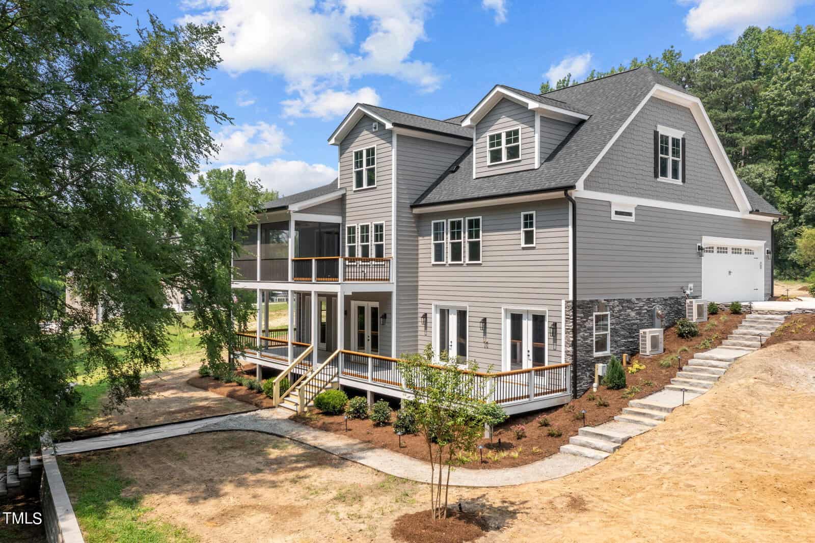 A large modern farmhouse-style home with gray siding, white trim, and stone accents. The house features multiple gables, a wraparound porch, large windows, and a landscaped yard under a blue sky with scattered clouds.