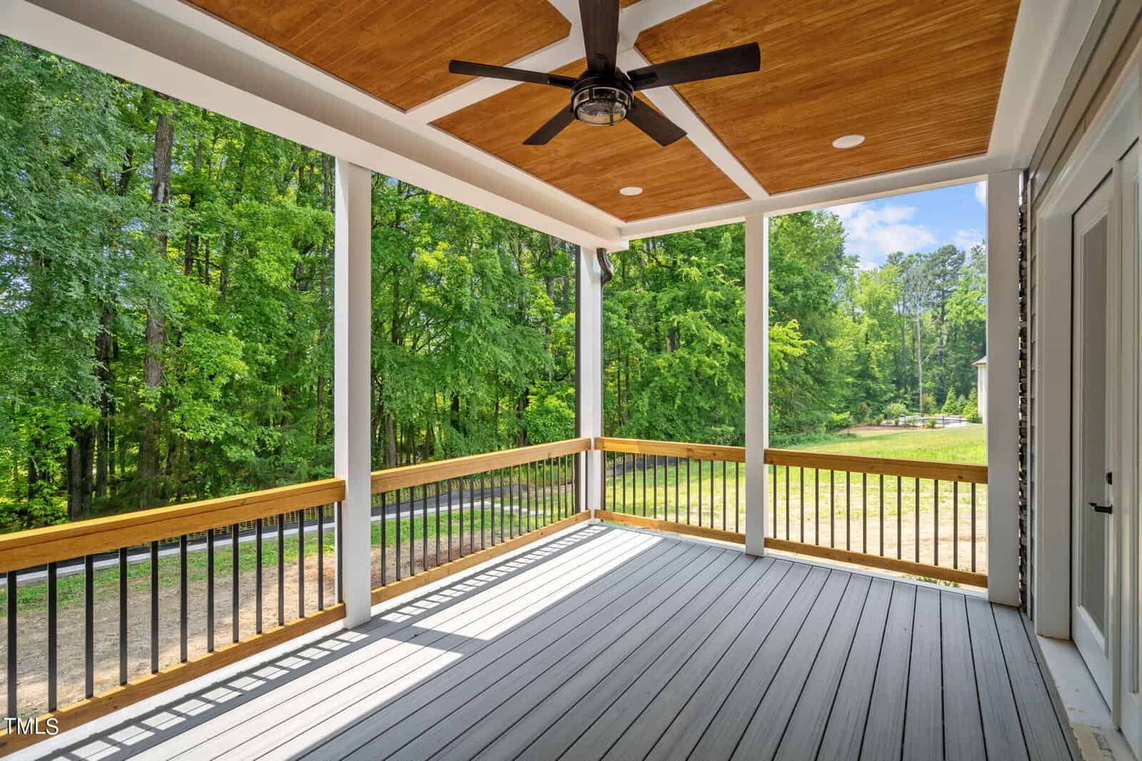 Covered porch with a ceiling fan, wood-paneled ceiling, white posts, and black railings, overlooking a green, wooded yard and a quiet driveway on a sunny day.