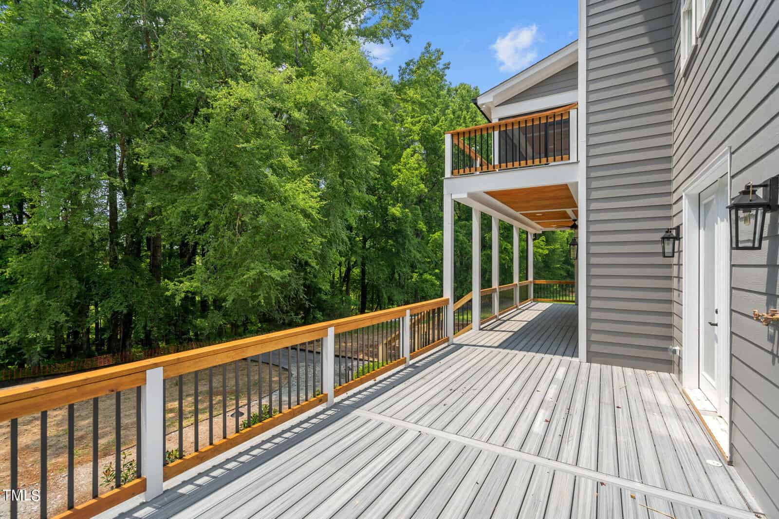 Spacious wooden deck with gray planks, wooden and metal railing, attached to a gray house; overlooks a wooded area and driveway under a clear blue sky.