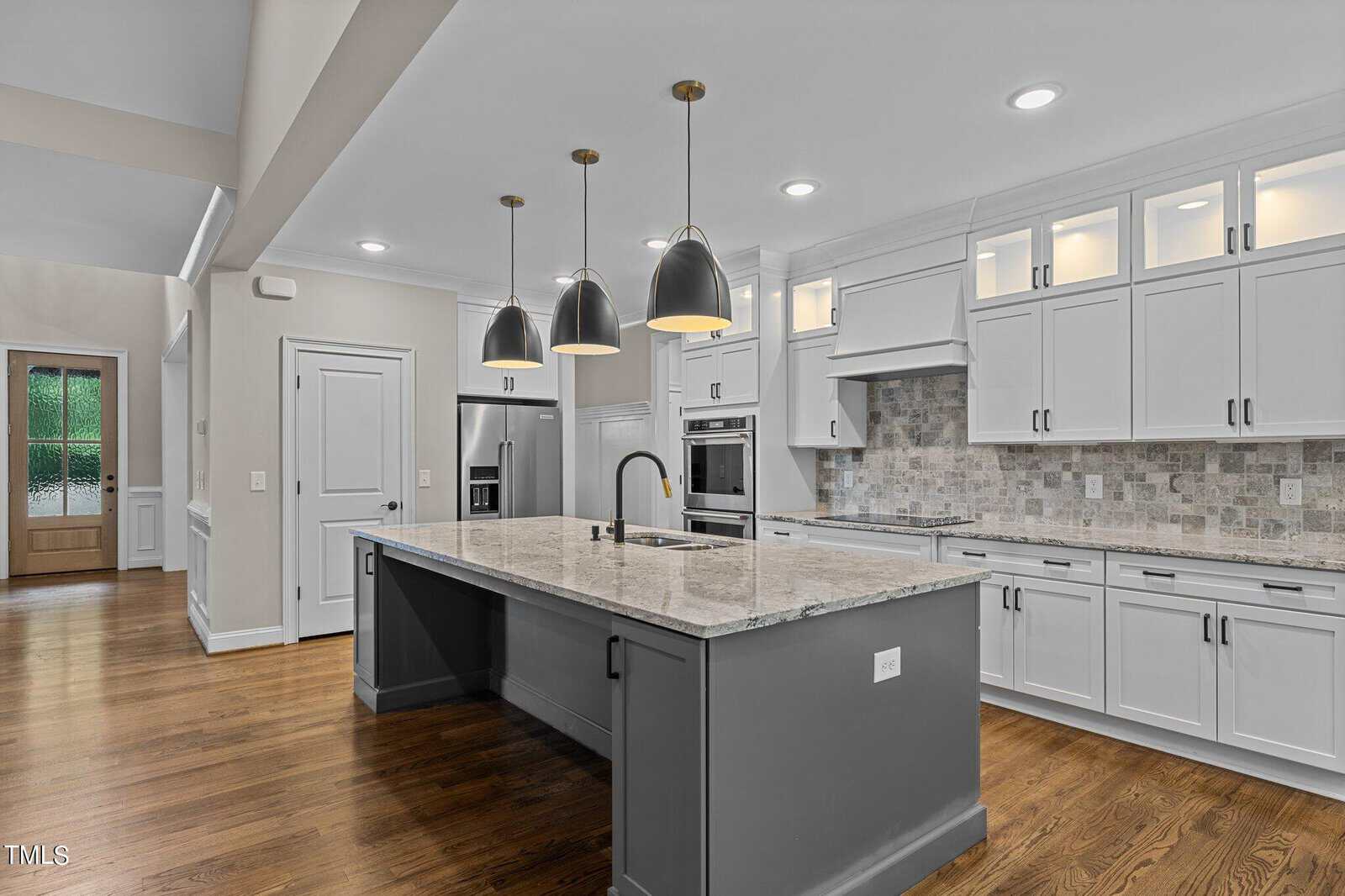 Modern kitchen with white cabinets, a large gray island with a marble countertop, three pendant lights, stainless steel appliances, wood flooring, and a tile backsplash. Door and natural light visible in the background.