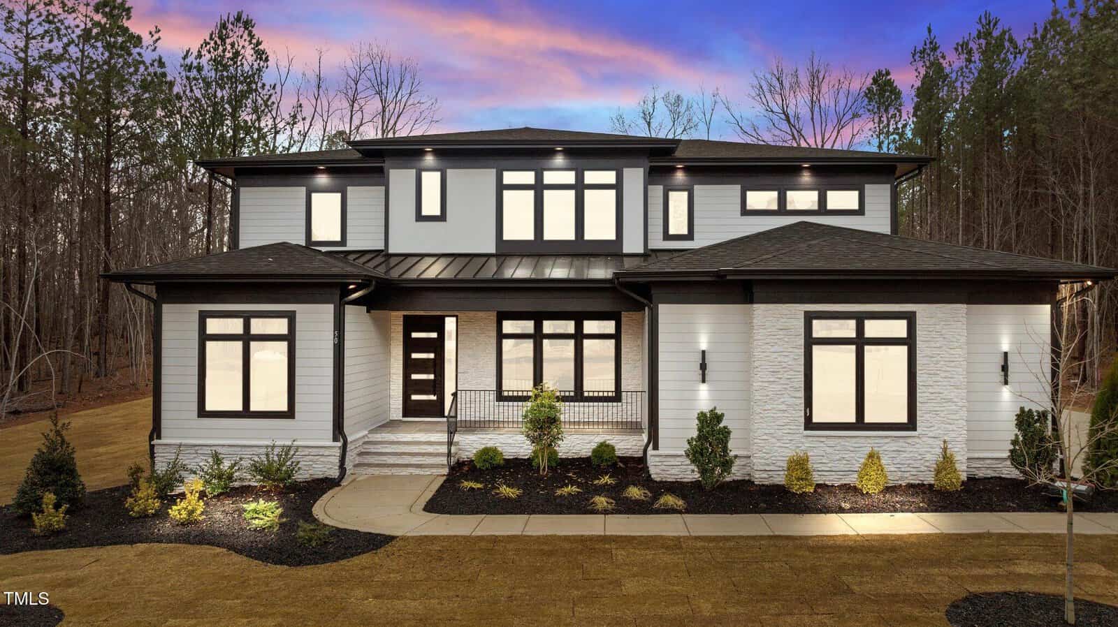 A modern two-story house with large windows, white siding, and a dark roof. The house is surrounded by a simple landscaped yard with small shrubs, and trees are visible in the background at dusk.