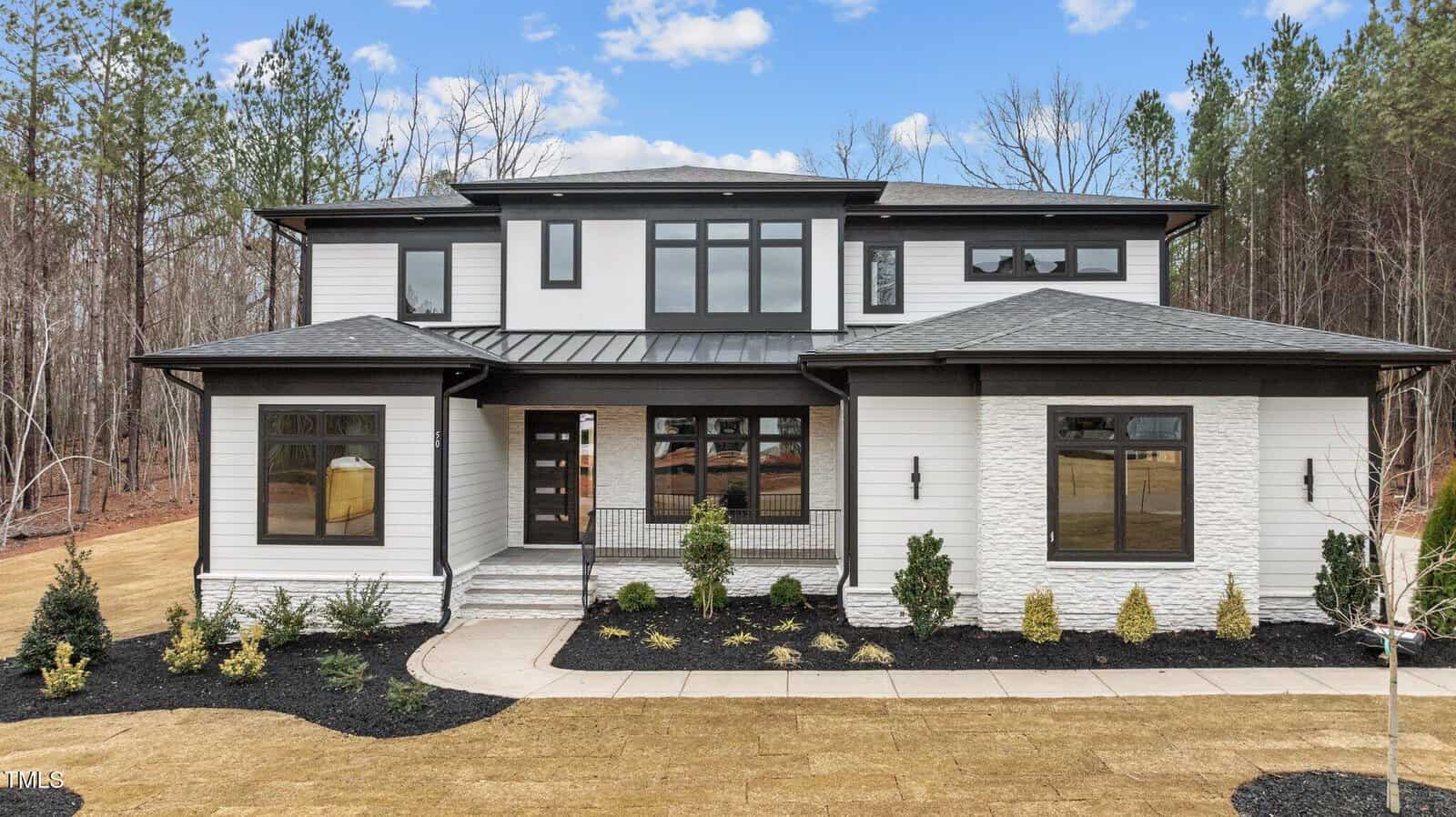 A modern two-story house with white siding, black trim, large windows, stone accents, and a neatly landscaped front yard, set against a backdrop of trees under a partly cloudy sky.