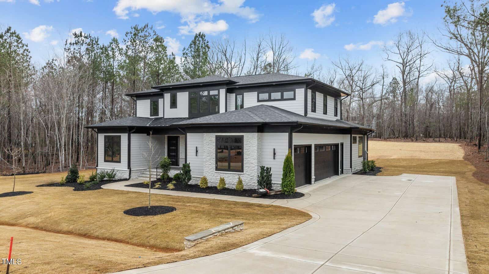 A two-story modern house with light gray siding, black trim, and a three-car garage, set on a large lot with a circular driveway, minimal landscaping, and trees in the background under a partly cloudy sky.