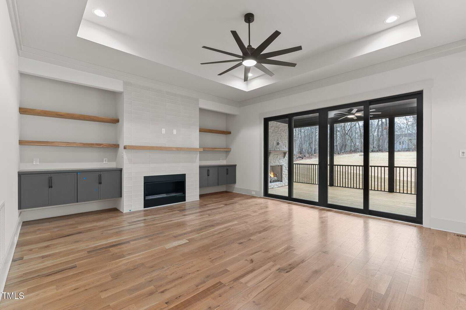 Spacious, modern living room with hardwood floors, a white tile fireplace, built-in shelves and cabinets, large sliding glass doors, and a view of a wooden deck and yard. Black ceiling fan in the center.