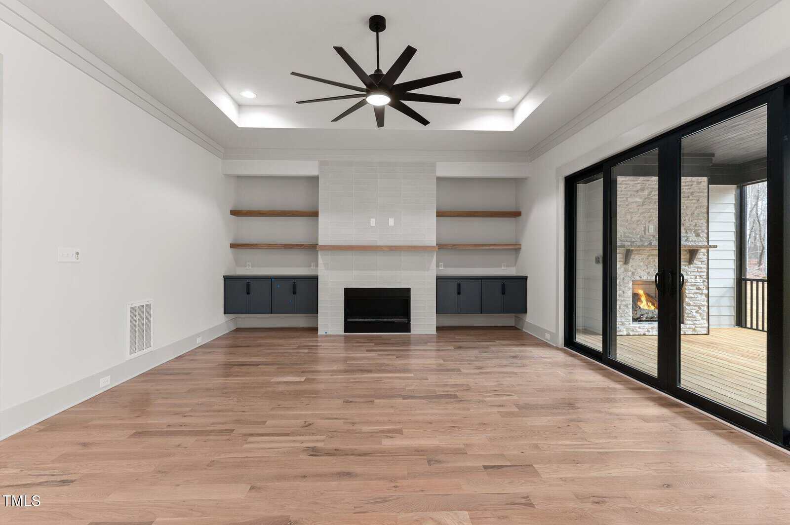 Modern living room with light wood floors, built-in shelves around a fireplace, dark cabinets, a large black ceiling fan, and sliding glass doors opening to a patio with an outdoor fireplace.