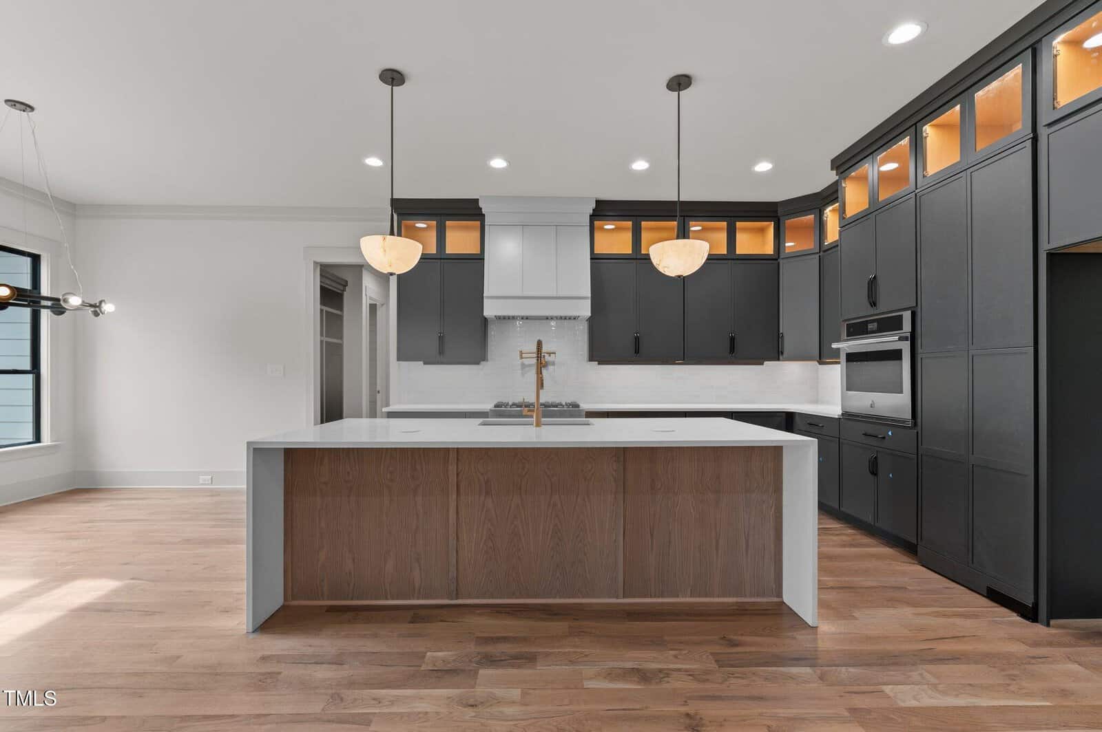 Modern kitchen with black cabinets, white countertops, a large island with a wood accent, double wall ovens, pendant lights, under-cabinet lighting, and light wood flooring.