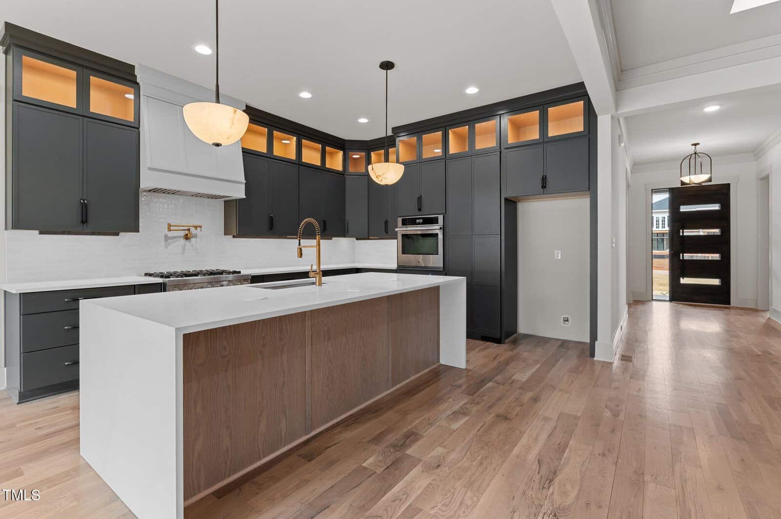 Modern kitchen with dark cabinets, light countertops, a large island with a wood base, built-in oven, gas stove, gold fixtures, pendant lights, wood flooring, and an open view to the hallway and front door.