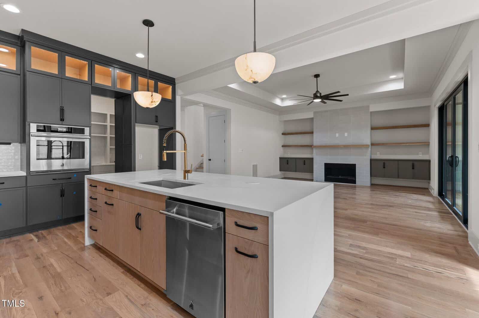 Modern kitchen with a large island featuring a sink and dishwasher, pendant lights overhead, built-in oven, gray cabinets, open shelves, and an adjacent living area with a fireplace and wall shelving.