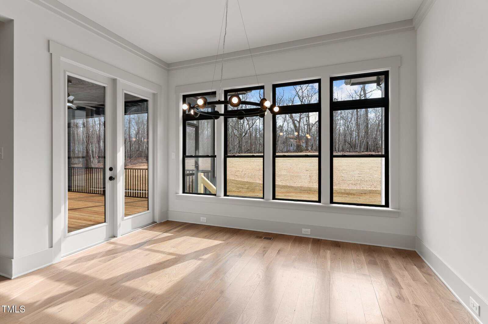 A bright, empty room with light wood flooring, large black-framed windows overlooking a grassy yard and trees, a modern black chandelier, and a glass door leading to a covered deck.