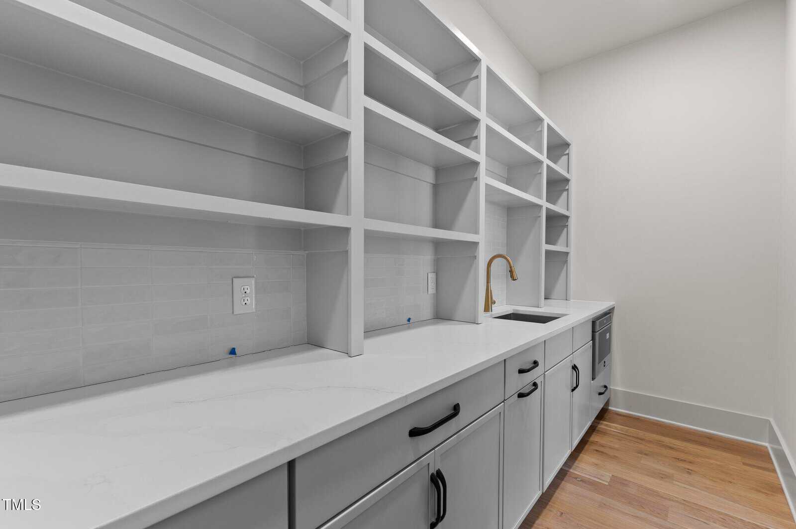 A modern pantry with white open shelving, white countertops, a stainless steel sink with a gold faucet, light gray cabinets, and light wood flooring. White subway tile backsplash and electrical outlets are visible.