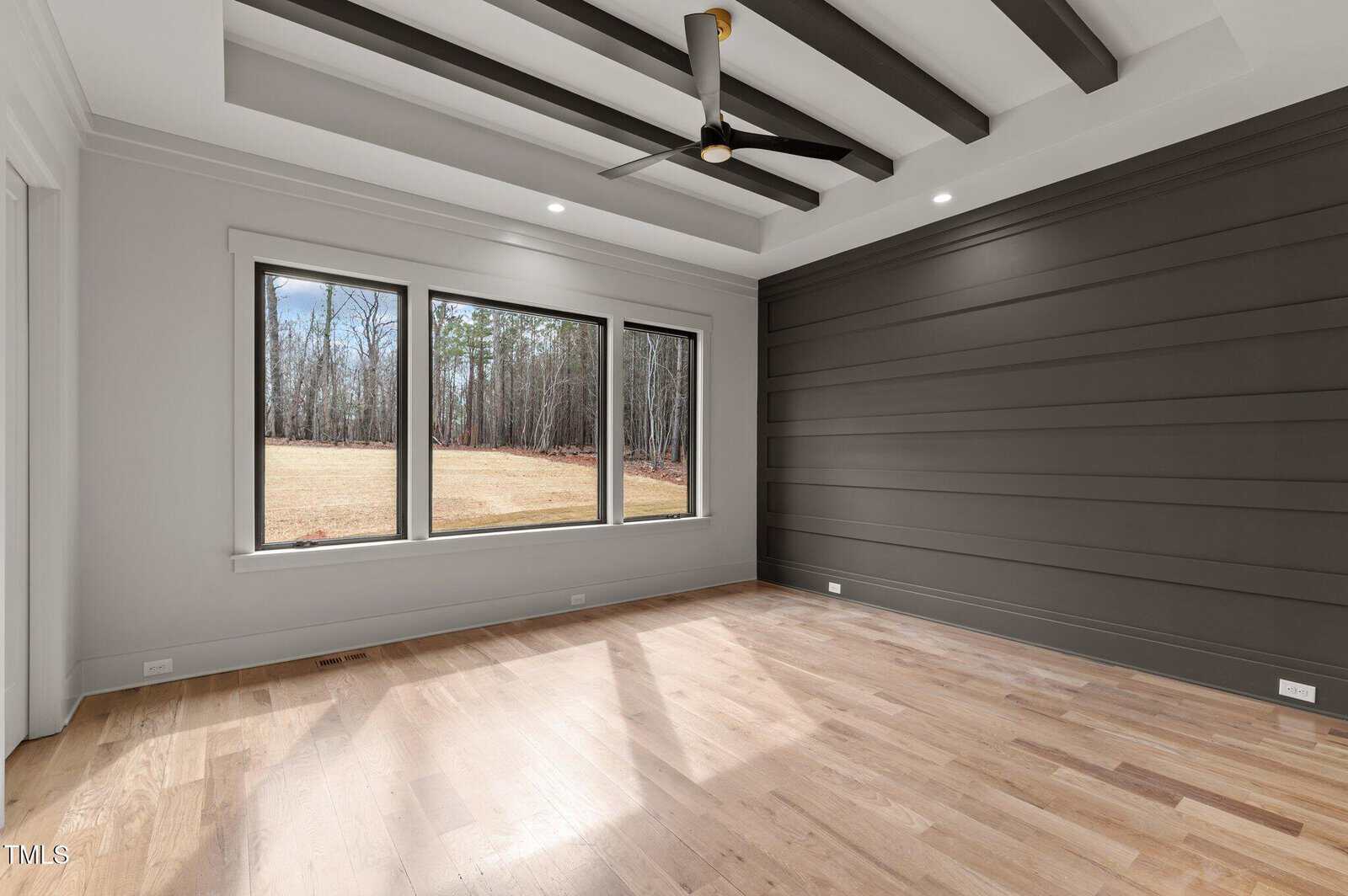 Modern empty room with wood floors, large windows showing a grassy yard and trees, a dark accent wall with horizontal paneling, and a ceiling fan with exposed beams.
