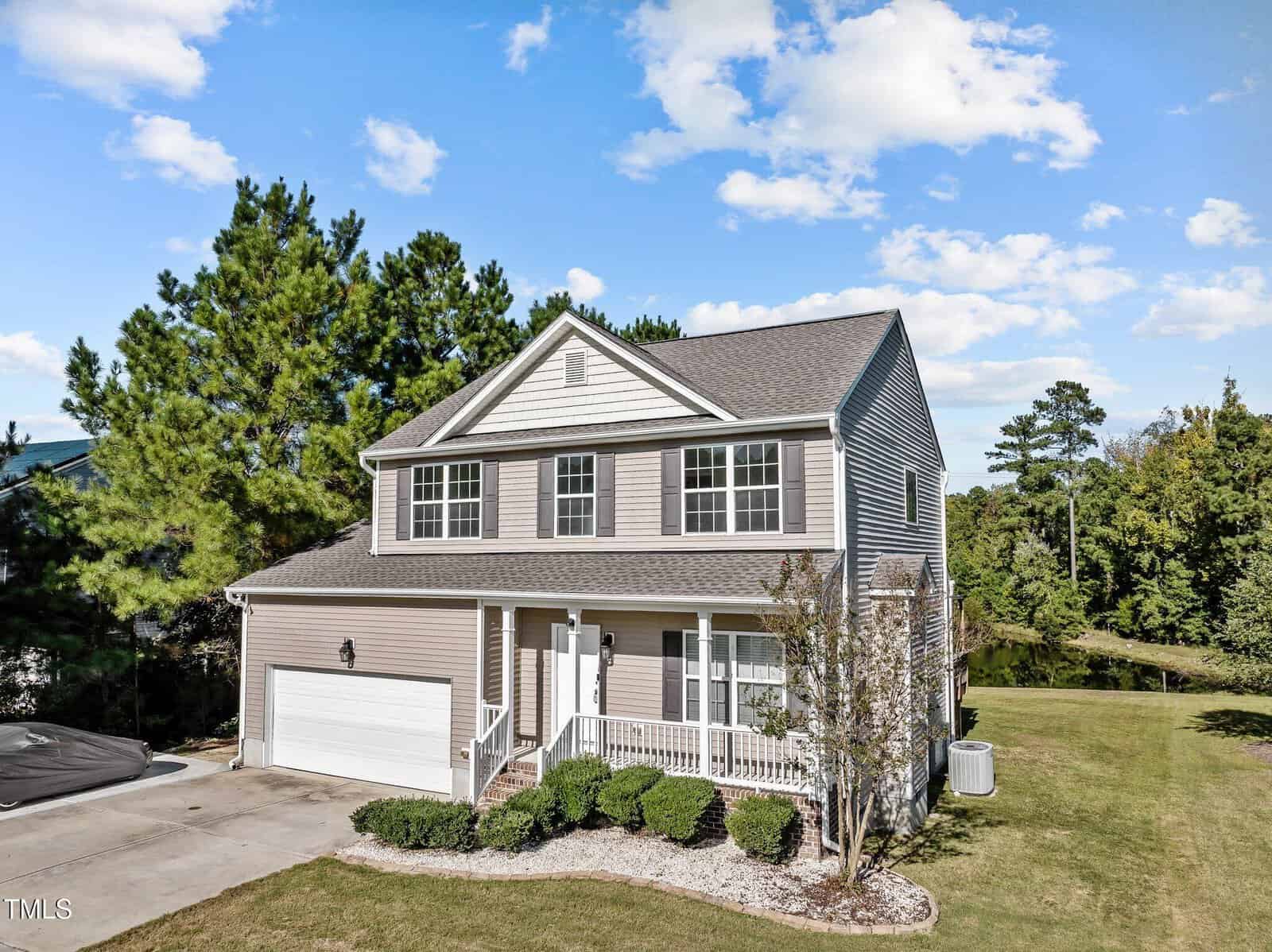 A two-story house with a front porch, light brown siding, and a two-car garage. It's surrounded by a well-maintained lawn and trees under a blue sky with scattered clouds.