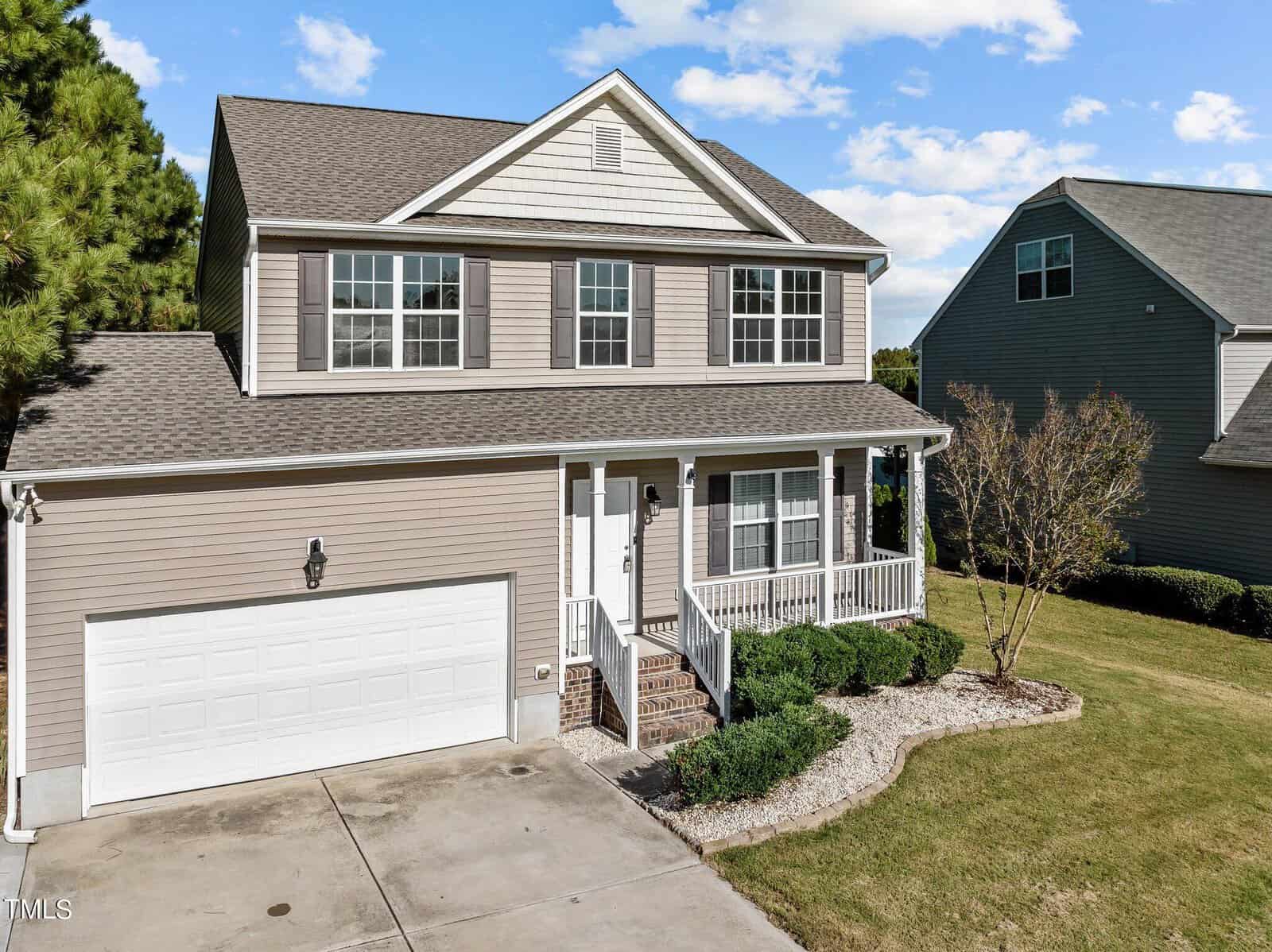 A two-story house with beige siding and a white garage door. The front features a small porch with white railings, shrubs lining the walkway, and a neatly trimmed lawn. Another house is visible in the background under a blue sky with clouds.