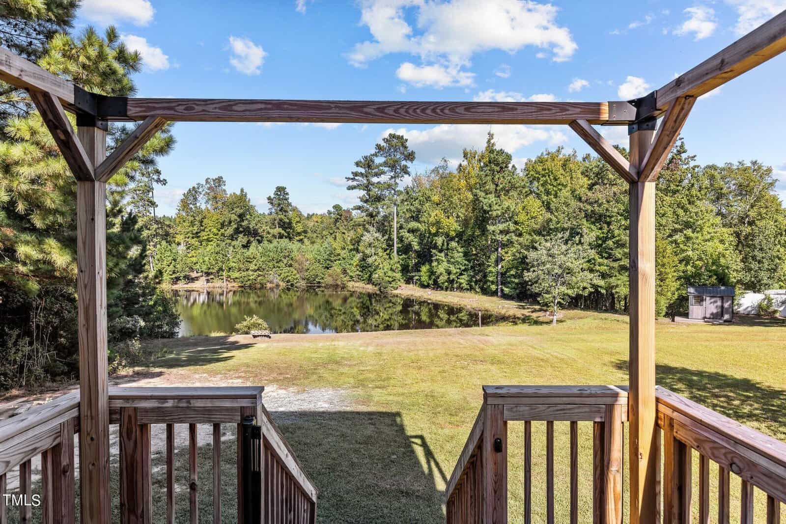 Wooden stairs lead down to a grassy area with a small pond surrounded by trees. The sky is mostly clear with a few clouds, creating a peaceful, natural setting.