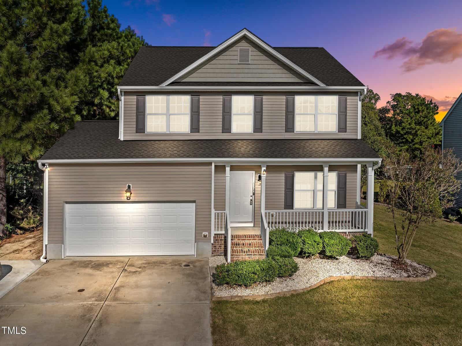 Situated at 723 Forrest Lane, this two-story gray house in Creedmoor boasts a charming covered porch. It features a double garage and a well-maintained front yard with shrubs and a small tree. The sky is a gradient of blue and orange, suggesting evening or early morning.