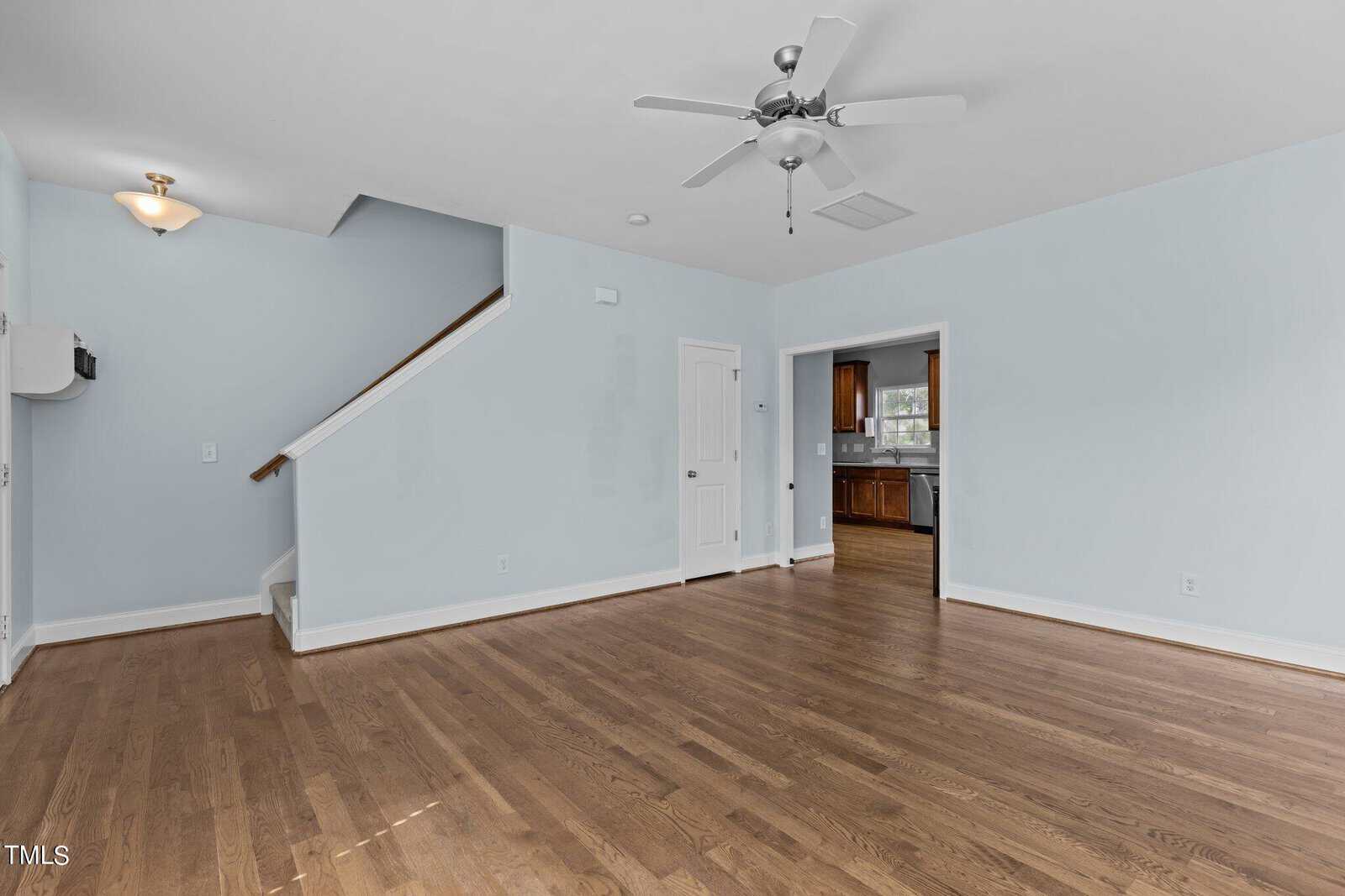 A spacious living room with light blue walls, hardwood floors, and a ceiling fan. An open doorway leads to a kitchen with wooden cabinets. A staircase is visible on the left side, and a small light fixture is on the ceiling.
