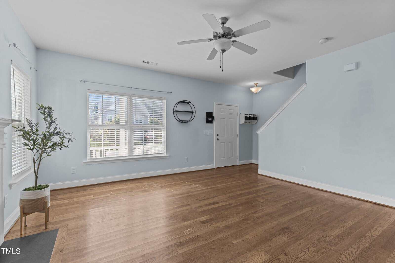 A spacious empty living room with light blue walls and hardwood floors. Features a ceiling fan, large window with blinds, a potted plant, a wall mirror, and a staircase leading to an upper floor. Natural light brightens the room.