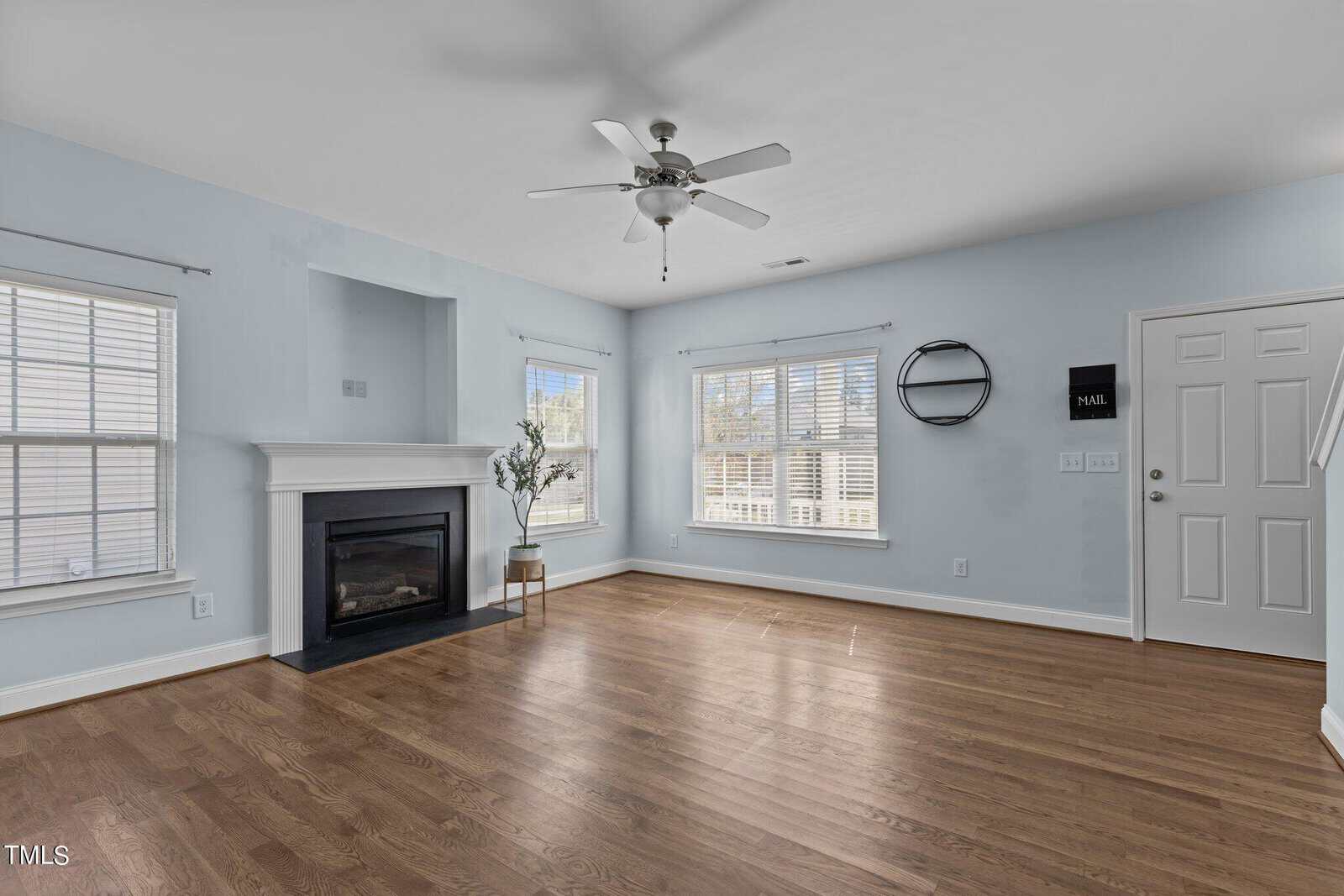 A spacious living room with light blue walls and hardwood flooring. It features a ceiling fan, a fireplace, large windows with blinds, and a circular wall decor piece. There is a plant near the fireplace and a white door on the right.