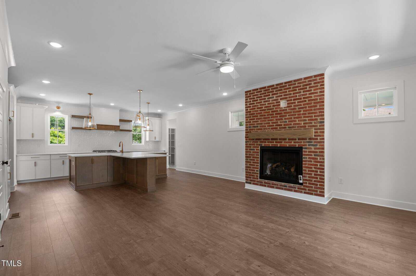 Open-concept living area with wood flooring, a brick fireplace, white walls, and recessed lighting. The kitchen features an island with pendant lights, white cabinets, and two windows providing natural light.