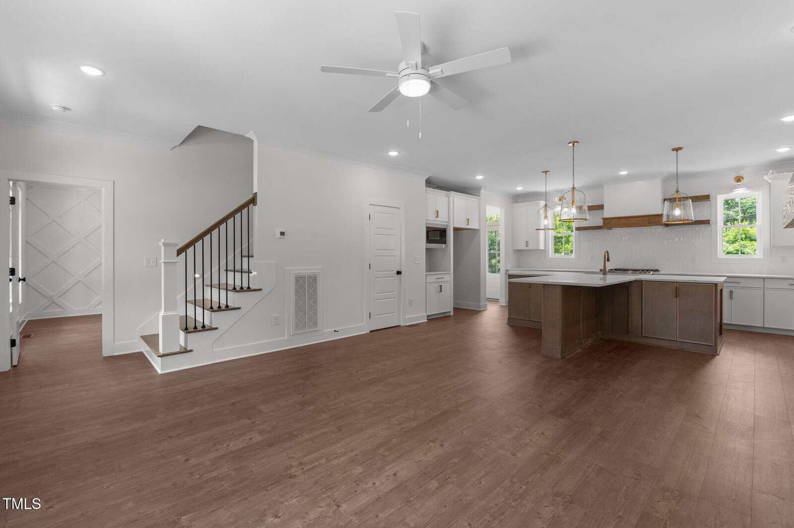 A spacious, modern open-concept kitchen and living area features wood flooring, white walls, built-in cabinets, a center island, pendant lights, a ceiling fan, and a staircase leading to the upper floor.
