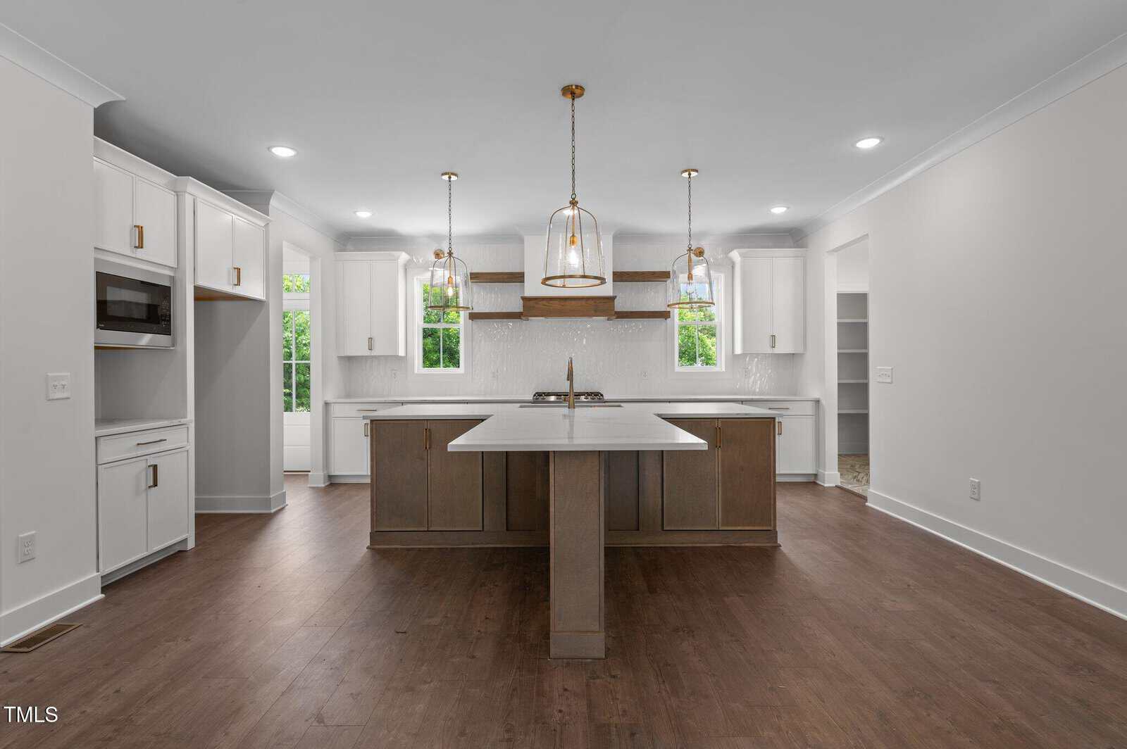 A modern kitchen with white cabinets, a center island with brown wood base, three pendant lights, built-in microwave, and large windows letting in natural light. The floor is dark wood and the walls are light gray.