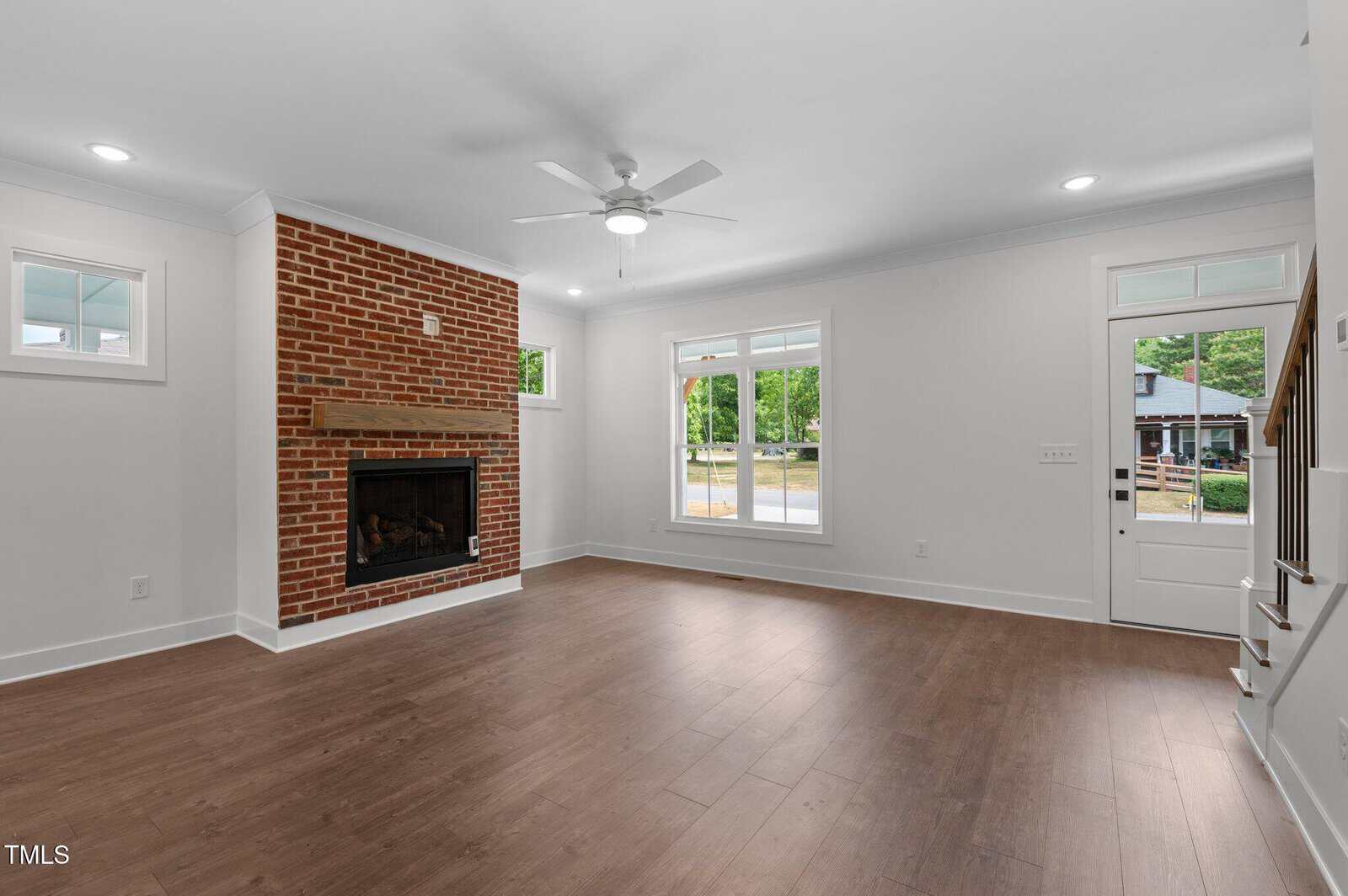 A spacious living room with wood flooring, white walls, a brick fireplace with a wooden mantle, large windows, a ceiling fan, and a door leading outside. Stairs are visible on the right.