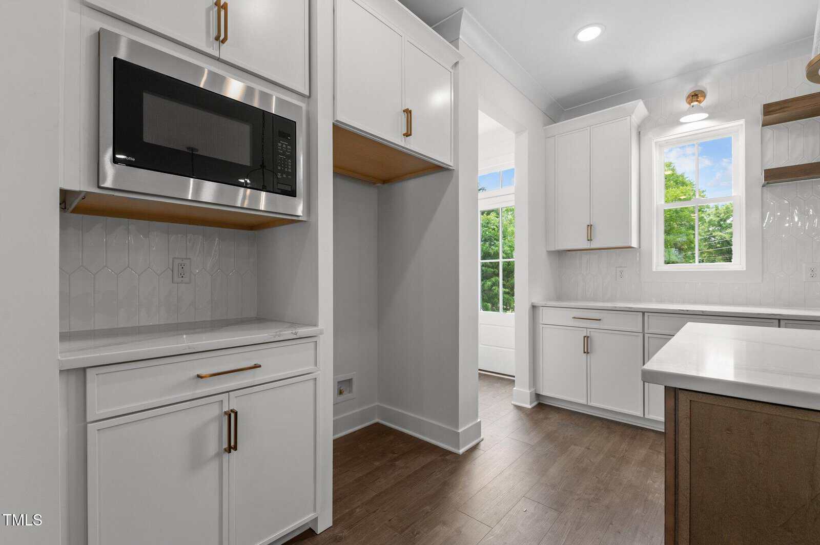 Modern kitchen with white cabinets, gold hardware, a built-in microwave, wood flooring, open shelving, and a window letting in natural light. The countertops and backsplash are white, creating a clean and bright look.