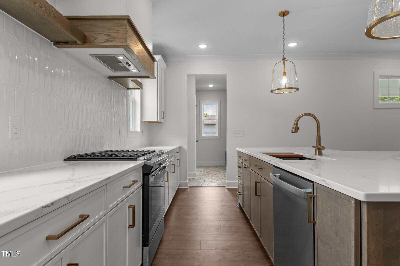 Modern kitchen with white cabinets, marble countertops, stainless steel appliances, gold hardware, a central island with a built-in sink, pendant lights, and wood flooring. A doorway leads to another room in the background.