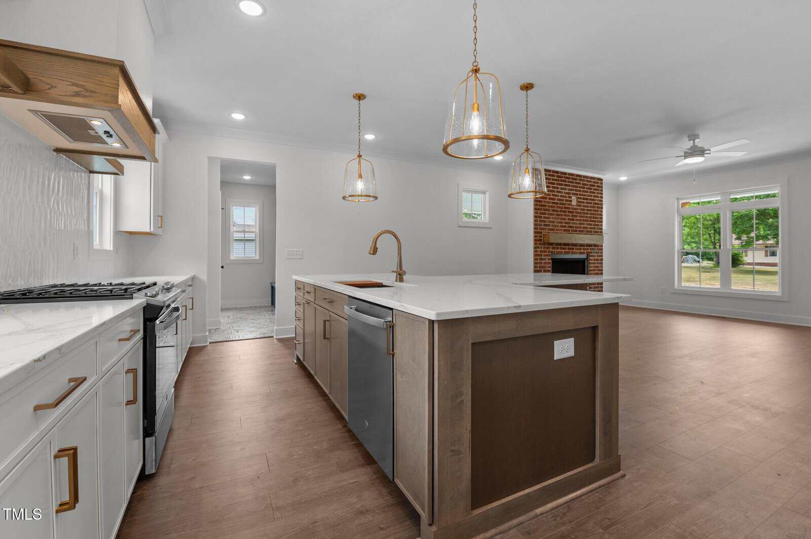 Modern open-concept kitchen and living area with a large island, pendant lights, stainless steel appliances, white cabinets, wood floors, brick fireplace, and windows showing a green outdoor view.