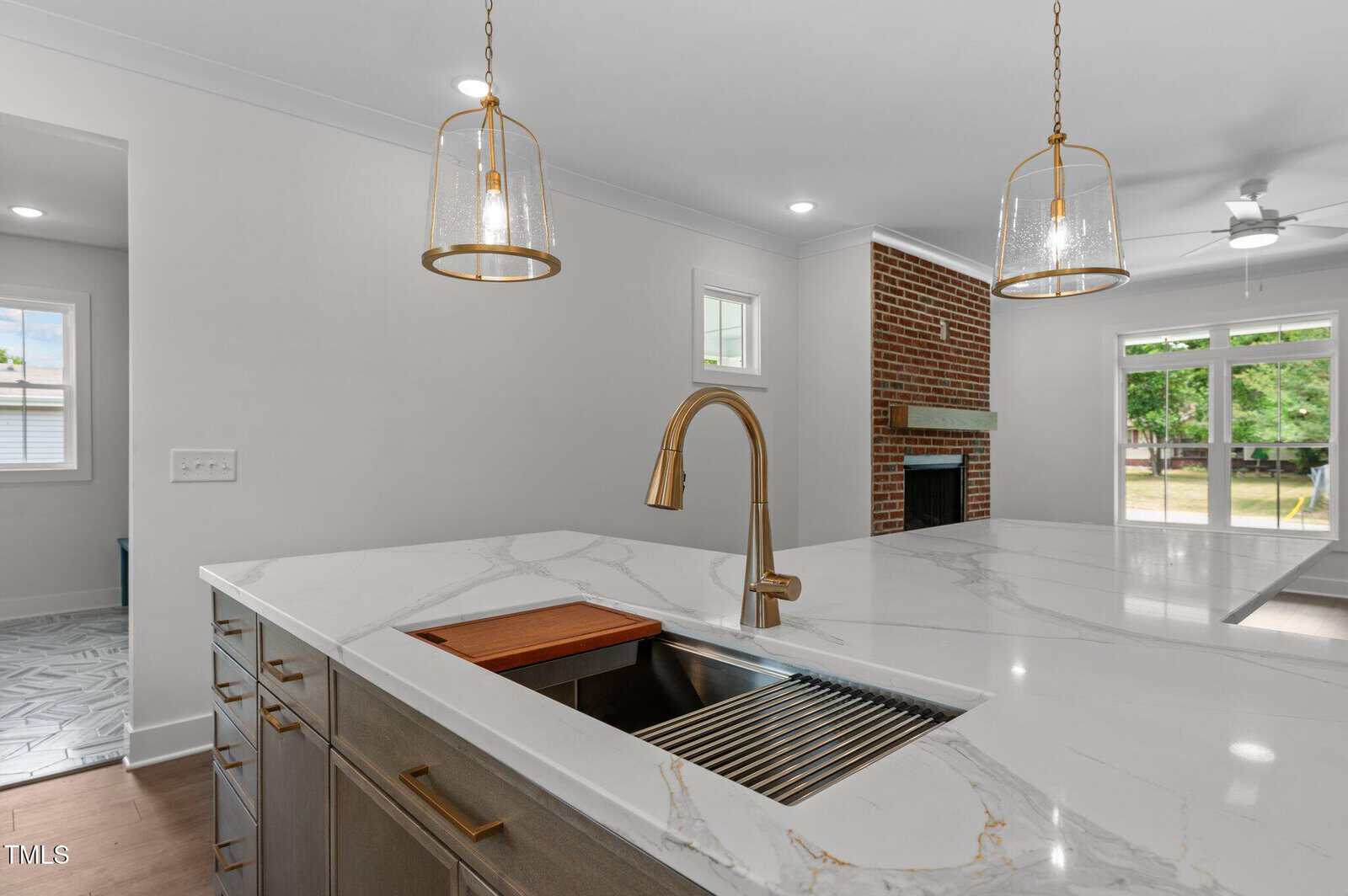 A modern kitchen island with a marble countertop, undermount sink, gold faucet, and built-in cutting board. Two pendant lights hang above. In the background, a brick fireplace and large windows are visible.