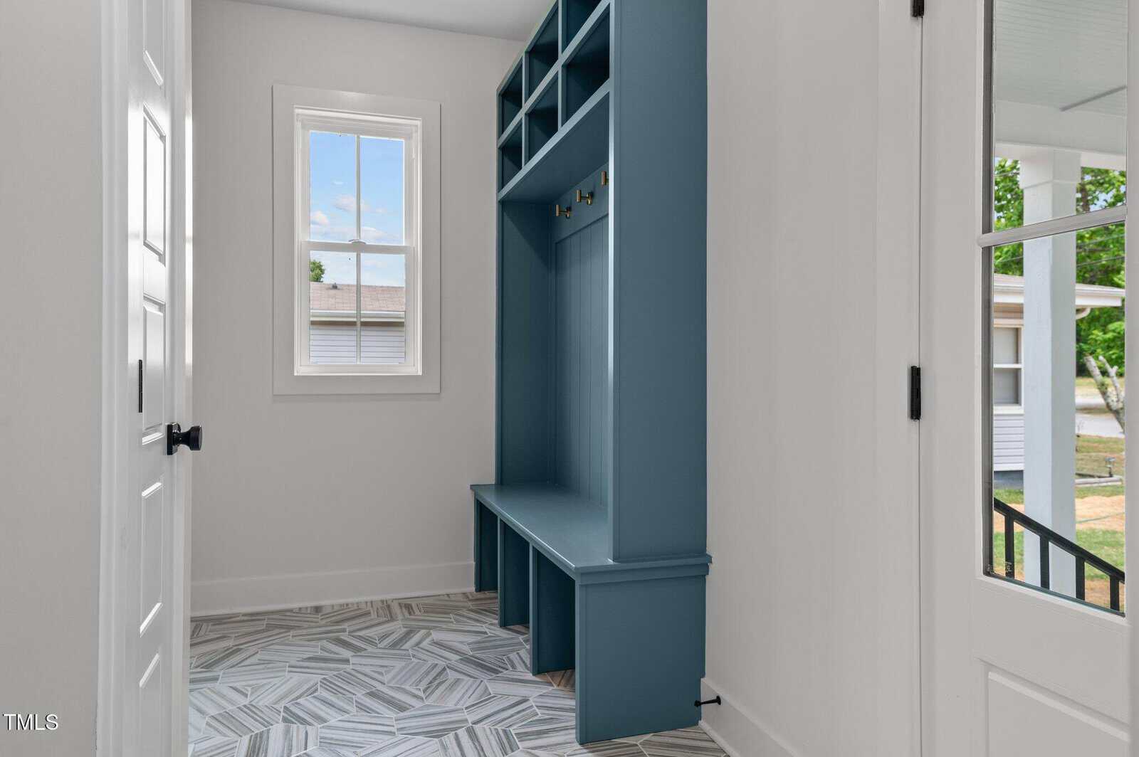A bright entryway with patterned tile floor, a blue built-in bench with cubbies and hooks, a window, and a door with glass panes leading outside.