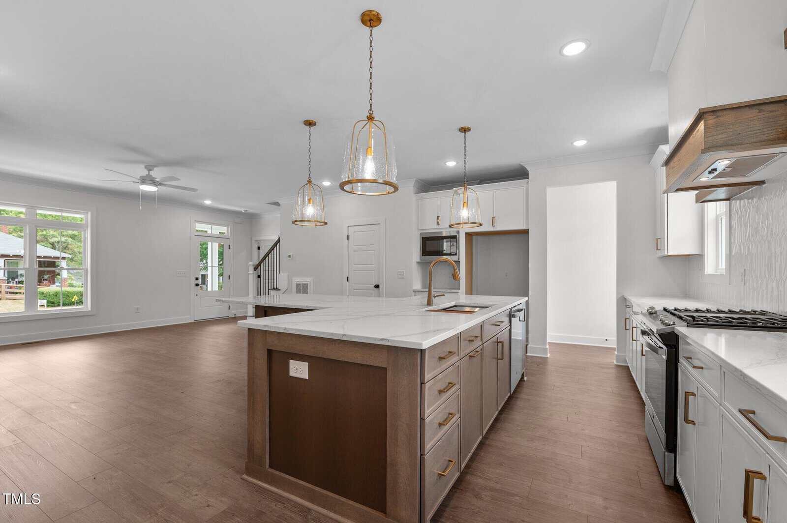 Spacious modern kitchen with an island, pendant lights, stainless steel appliances, white cabinets, gas stove, and hardwood flooring; open to a living area with large windows and a ceiling fan.