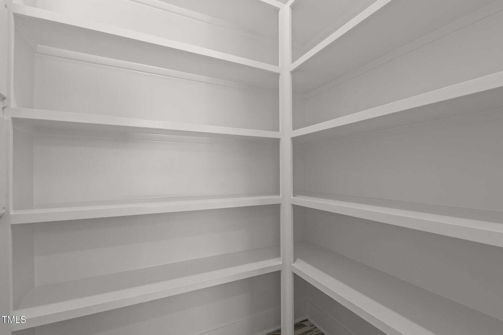 Empty walk-in pantry with white built-in shelves lining the walls. The shelves are clean and evenly spaced, creating ample storage space. The floor is visible at the bottom, and the overall space is well-lit.