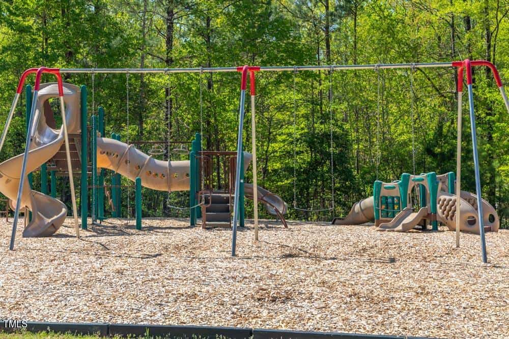 A playground with a spiral slide, climbing structures, swings, and wood chip ground cover, surrounded by green trees on a sunny day.