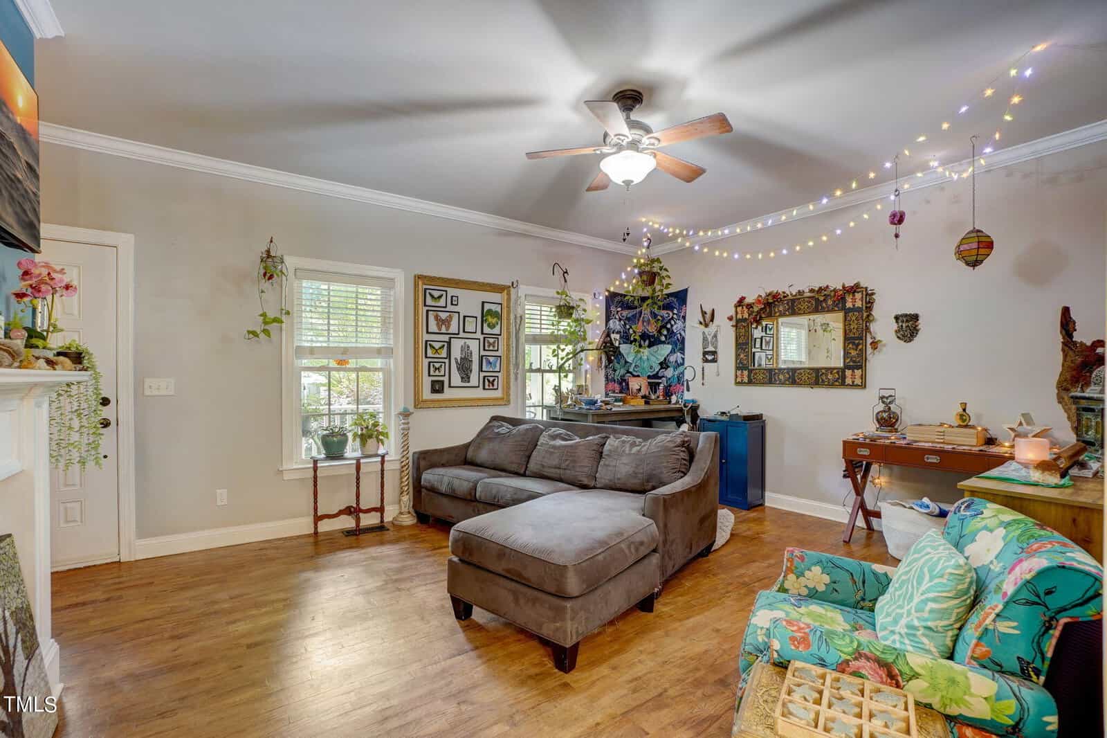 A cozy living room with hardwood floors, a gray sectional sofa, a floral armchair, a ceiling fan, string lights, wall decorations, plants, a mirror, and a small desk with a chair against the wall.