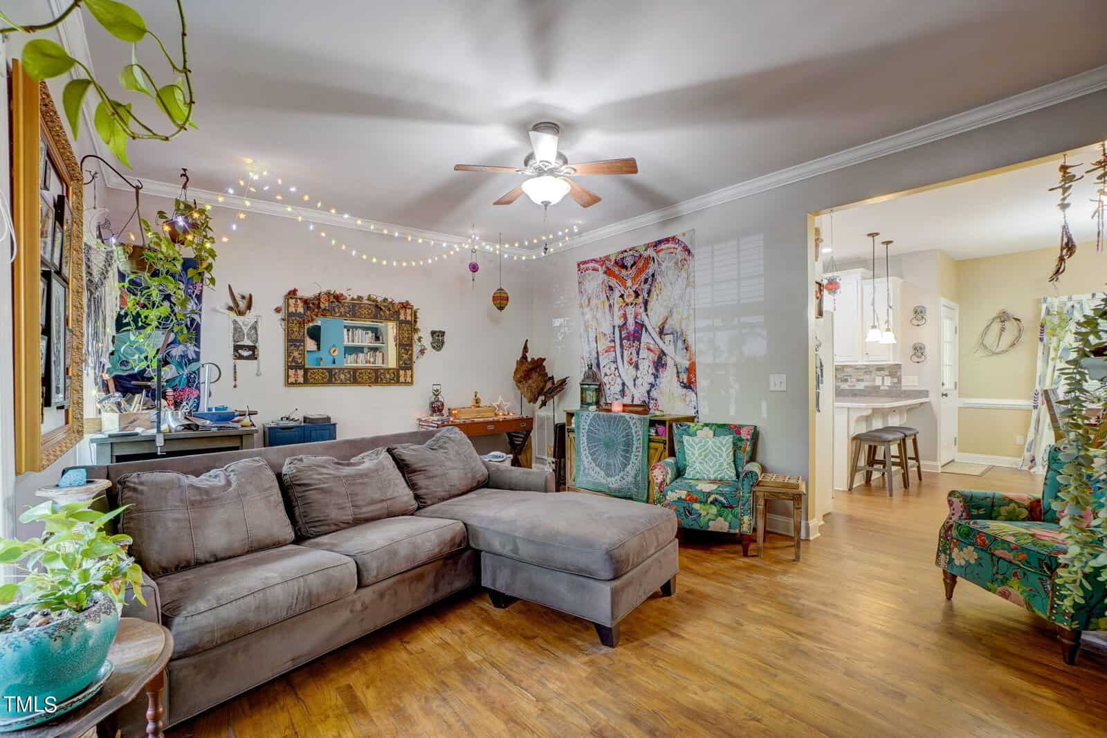 A living room with a gray sectional sofa, patterned armchair, and plants. Colorful tapestries and string lights decorate the walls. The space connects to a dining area and kitchen with wood floors throughout.