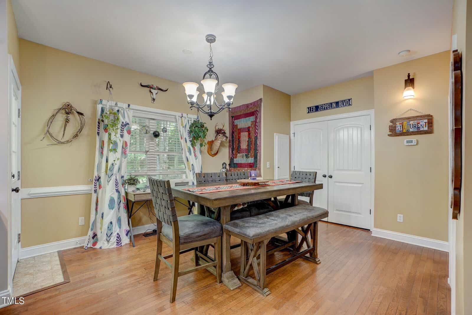 A dining room with tan walls, a wooden table and bench seating, a chandelier, hardwood floors, wall art, a window with blinds, and a double-door closet. The space is decorated with various rustic and western accents.