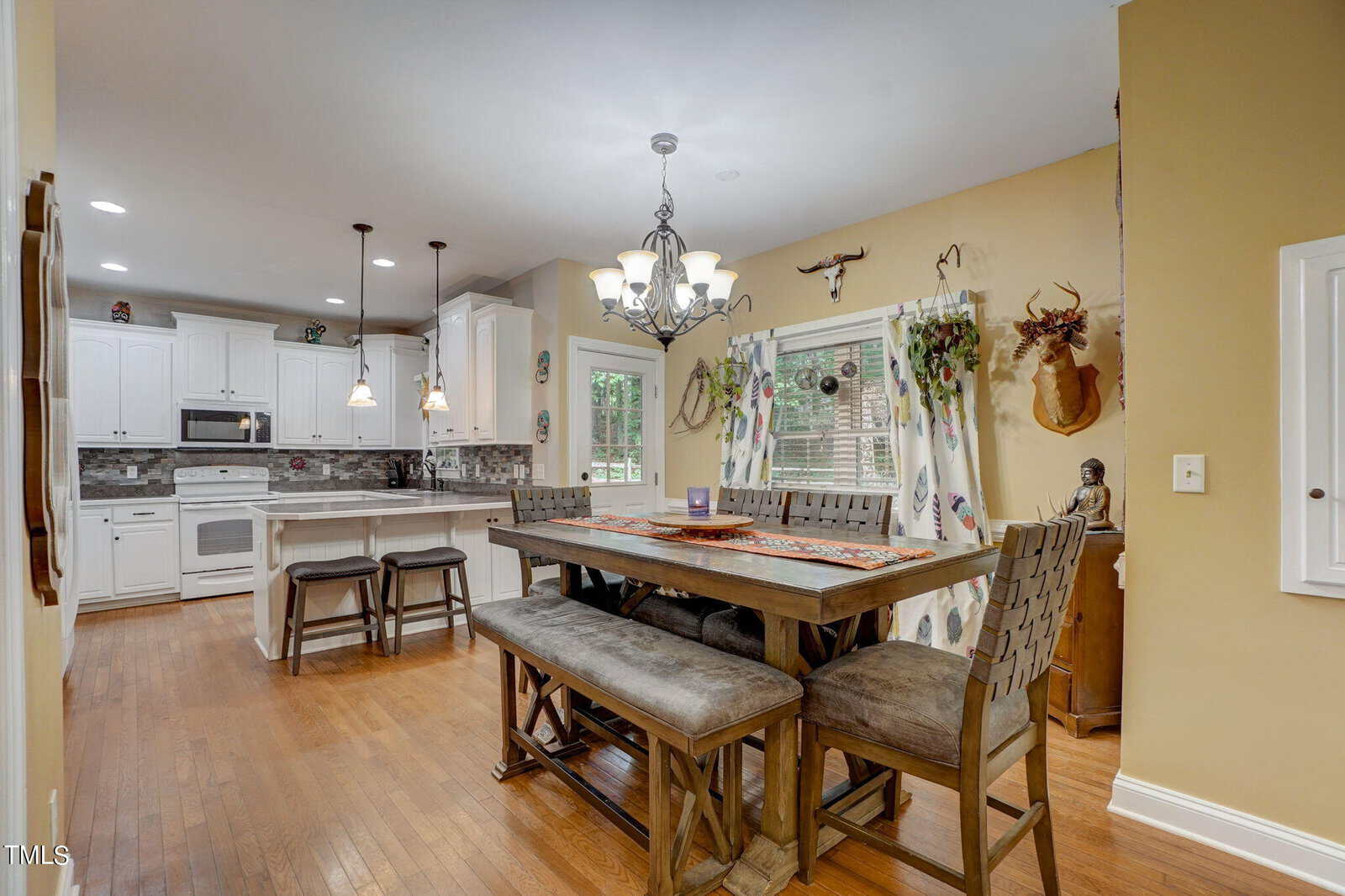 A kitchen and dining area with hardwood floors, white cabinets, an island with stools, and a wooden dining table with high-backed chairs. Wall decor and a window with patterned curtains are visible.