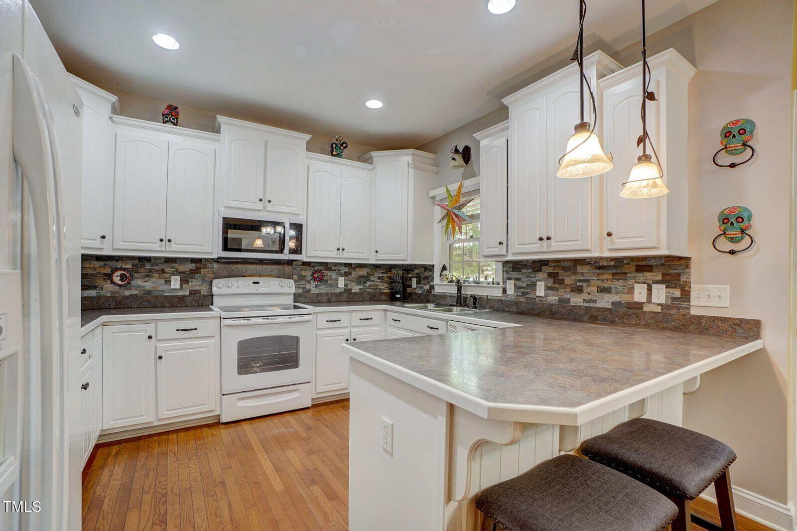 A kitchen with white cabinets, stainless steel appliances, a tile backsplash, wood floors, and two pendant lights over a gray countertop with an attached breakfast bar and two stools. Decorative items are on the cabinets and walls.