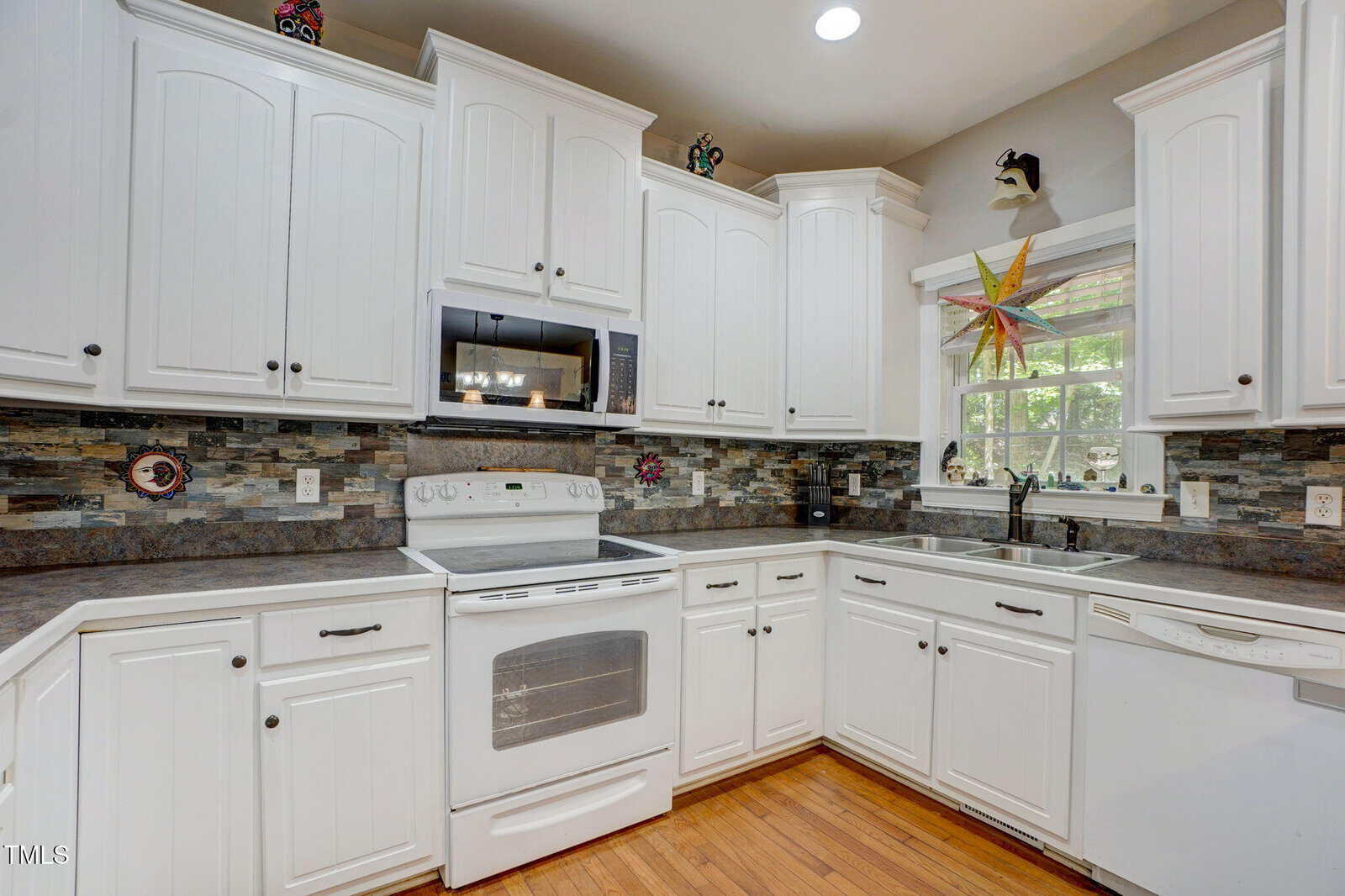 A kitchen with white cabinets, stainless steel appliances including an oven and microwave, a tile backsplash, a double sink under a window, and wood flooring. Decorative items are on top of the cabinets.